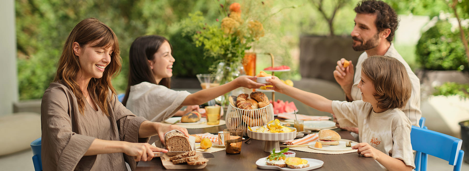 Eine Gruppe von vier Personen genießt ein gemeinsames Frühstück oder Brunch auf einer Terrasse. Die Personen - zwei Frauen, ein Mann und ein Kind - sitzen um einen dunklen Holztisch herum und lachen miteinander. Auf dem Tisch befindet sich eine reichhaltige Auswahl an Speisen: ein Korb mit Gebäck, ein Etagenständer mit Obst, Schüsseln mit Eierspeisen, verschiedene Brote, und Getränke wie Orangensaft. In der Mitte des Tisches steht ein großes Blumenarrangement mit grünen und orange-gelben Pflanzen. Die Szenerie spielt im Freien mit Pflanzen und Grünflächen im Hintergrund. Die Atmosphäre wirkt warm, freundlich und einladend.