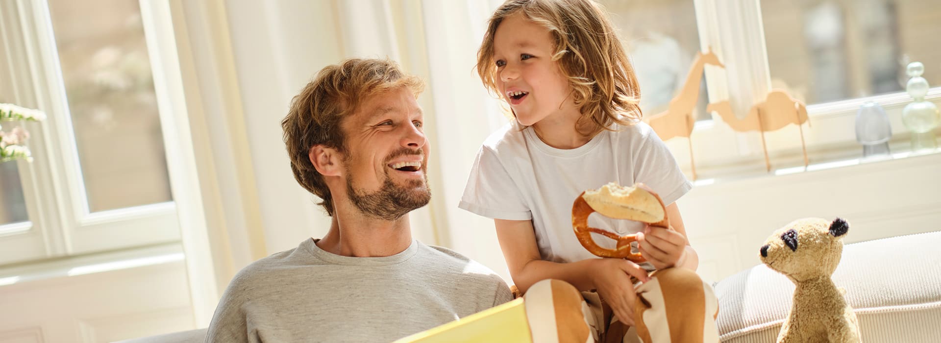 Ein lächelnder Mann mit kurzem, leicht gewelltem Haar und Bart sitzt auf einem Sofa und liest aus einem großen, gelben Buch vor. Neben ihm sitzt ein Kind mit schulterlangen, lockigen Haaren, das eine Brezel mit einem abgebissenen Stück in der Hand hält. Beide lachen und schauen sich an. Im Hintergrund sind helle Vorhänge, ein Fenster mit Holzspielzeug in Tierform auf der Fensterbank und ein Stofftier auf dem Sofa zu sehen.