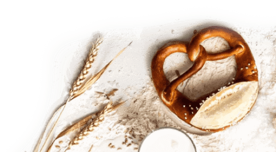 Eine Brezel liegt auf einer weißen, mit Mehl bestreuten Fläche. Links von der Brezel befinden sich zwei Getreideähren und verstreute Getreidekörner.