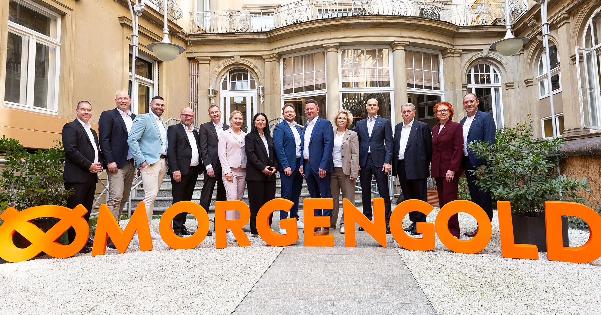 Eine Gruppe von 14 Personen in Business-Kleidung steht lächelnd in einem Innenhof. Im Vordergrund bilden große, orangefarbene 3D-Buchstaben und ein Logo den Schriftzug MORGENGOLD.