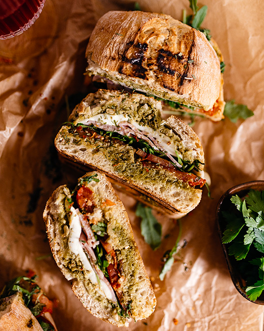 Italienisches Sandwich in Ciabatta-Brot, gefüllt mit Mozzarella, Tomaten, frischem Basilikum und grünem Pesto, auf braunem Papier serviert.