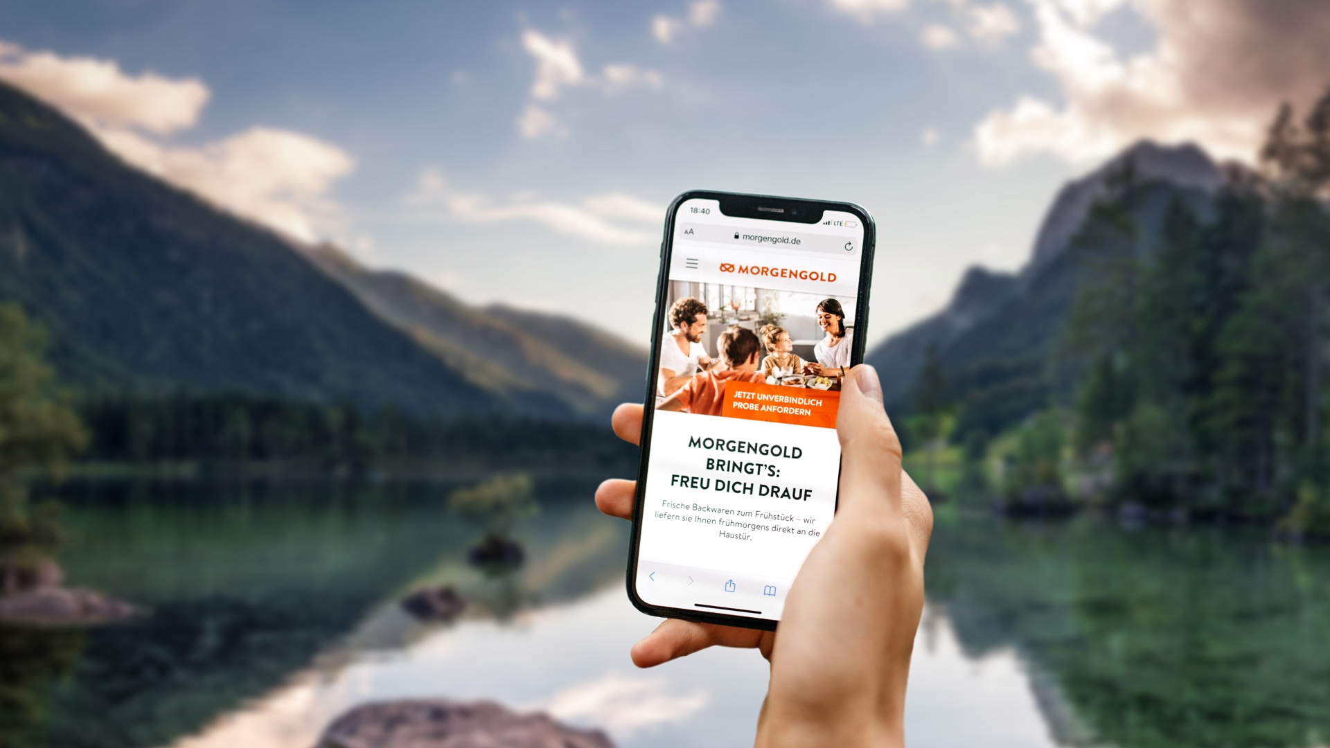 Hand hält Smartphone mit Morgengold-Website vor einem Bergsee in Deutschland.