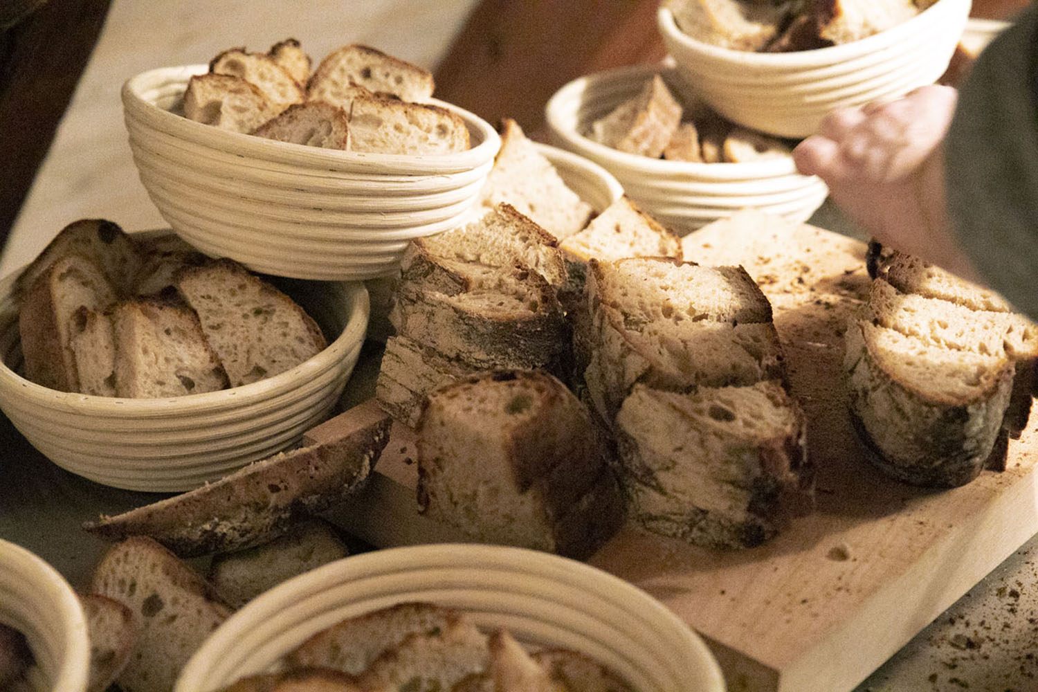 Eine Auswahl verschiedener Brotsorten in Körben und auf einem Holzbrett arrangiert, bereit für ein Brottasting. Im Vordergrund rustikale Brotscheiben, im Hintergrund greift eine Hand nach einer Scheibe.