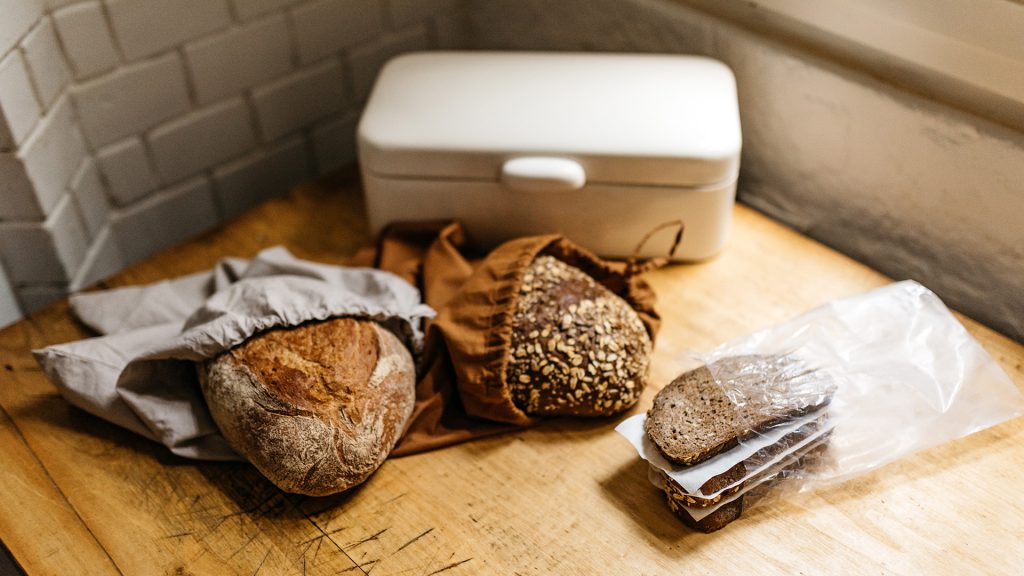 Verschiedene Brote in Stoffbeuteln und Plastik verpackt neben einem weißen Brotkasten auf einem Holztisch.