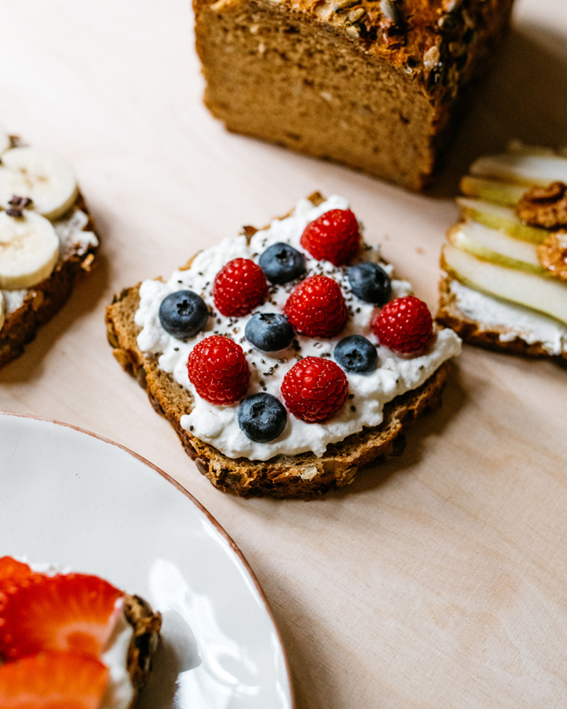 Scheibe Körnerbrot mit Frischkäse und Chiasamen, belegt mit frischen Himbeeren und Blaubeeren – daneben weitere Obstbrote und ein Laib Vollkornbrot.