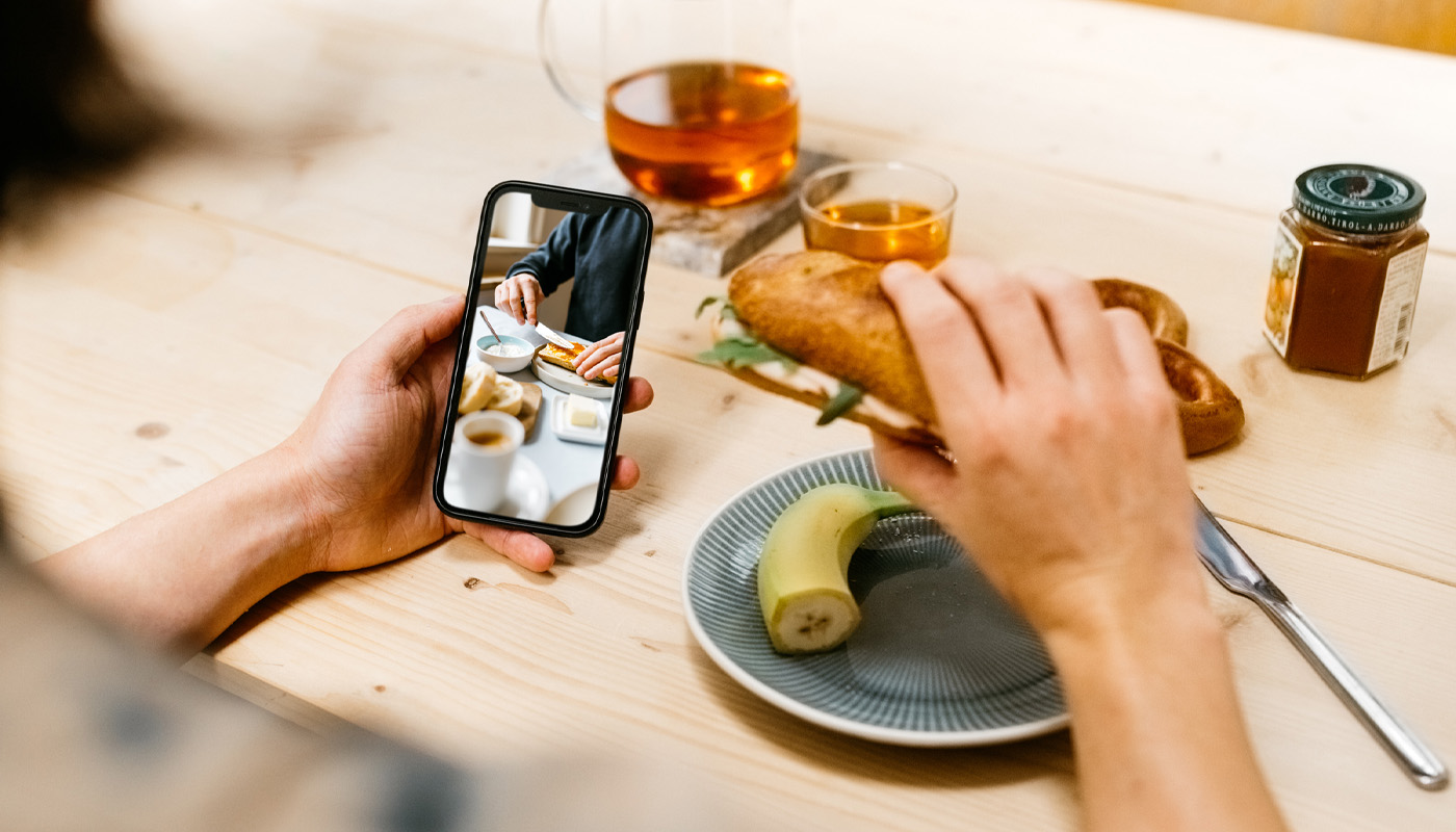 Person genießt ein belegtes Brötchen von Morgengold Frühstücksdienste und schaut dabei ein Frühstücksfoto auf dem Smartphone an, auf gedecktem Holztisch mit Tee, Banane und Marmelade.