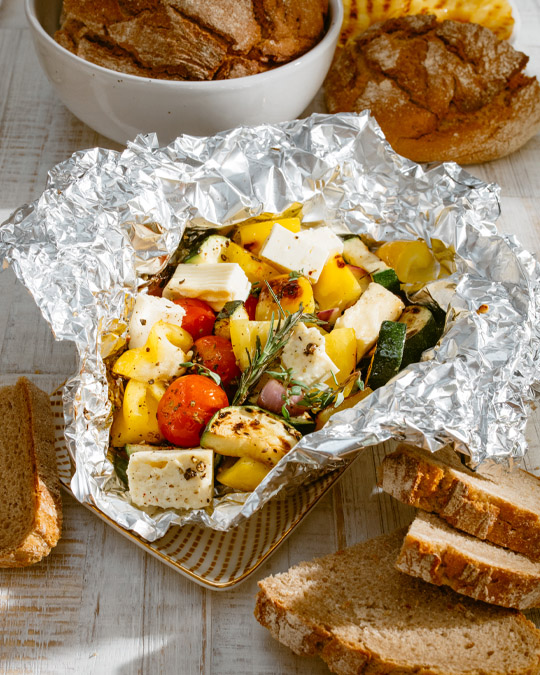 Grillgemüse mit Feta in Alufolie, serviert mit Brot und Brötchen auf hell gedecktem Tisch.
