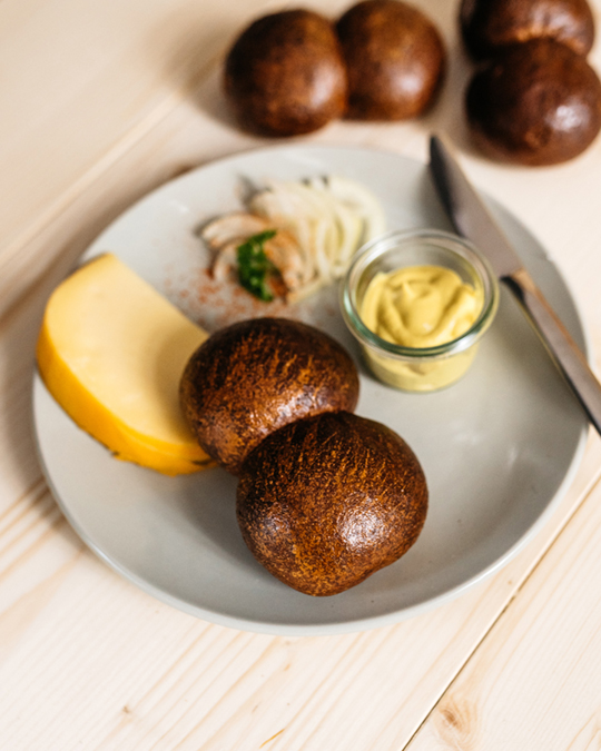 Zwei Röggelchen auf Teller mit Käse, Zwiebeln, Senf und Messer auf hellem Holztisch.