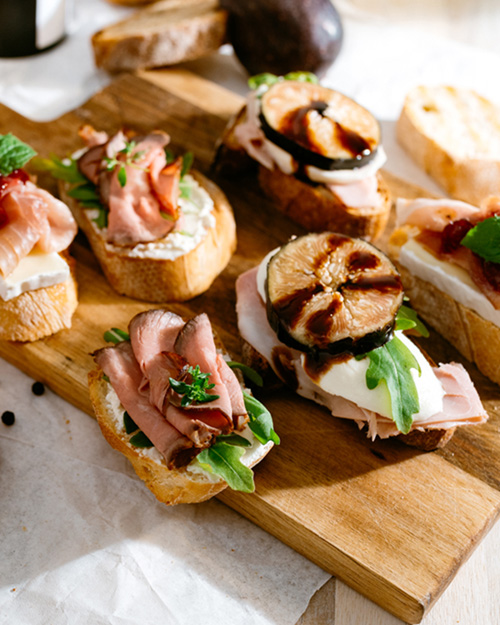 Frisch belegte Baguette-Scheiben mit Roastbeef, Rucola, Feigen und Frischkäse auf Holzbrett angerichtet.