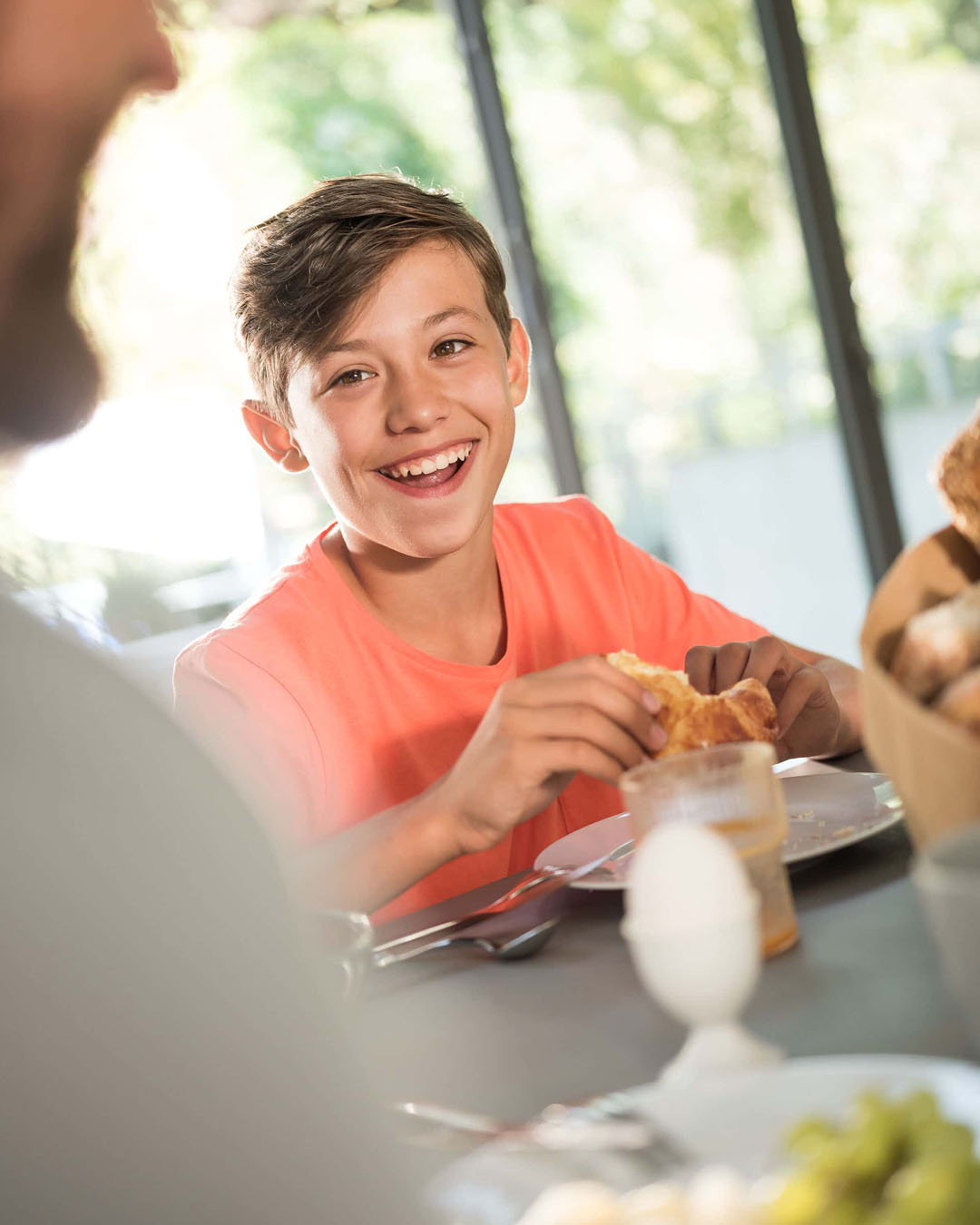 Junge lacht beim Frühstück und hält ein Croissant in den Händen, im Hintergrund gedeckter Tisch mit Frühstück.