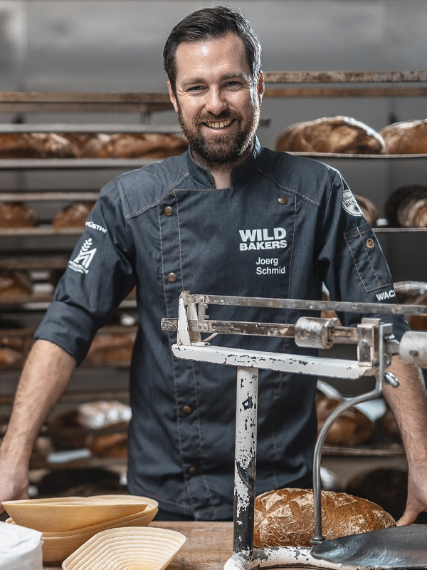 Meisterbäcker Jörg Schmid in der Backstube der Wildbakers – mit traditioneller Waage und frisch gebackenem Brot inmitten einer Handwerksbäckerei.