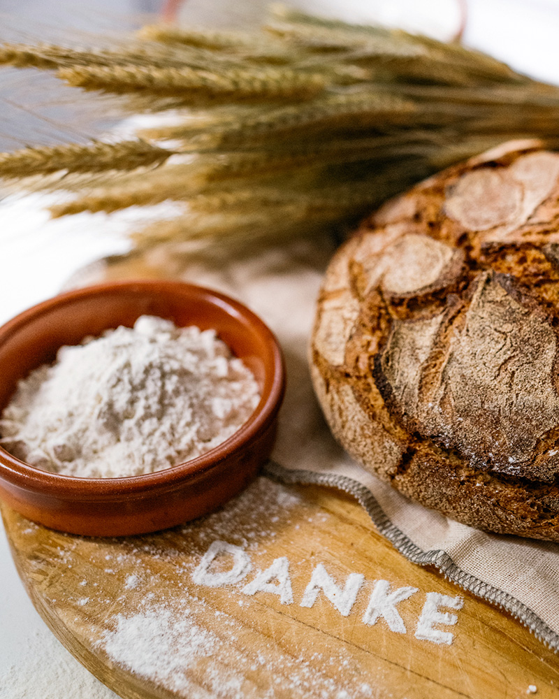 Rustikales Brotlaib mit Mehl und Ähren auf Holzbrett – das Wort „DANKE“ liebevoll mit Mehl geschrieben.