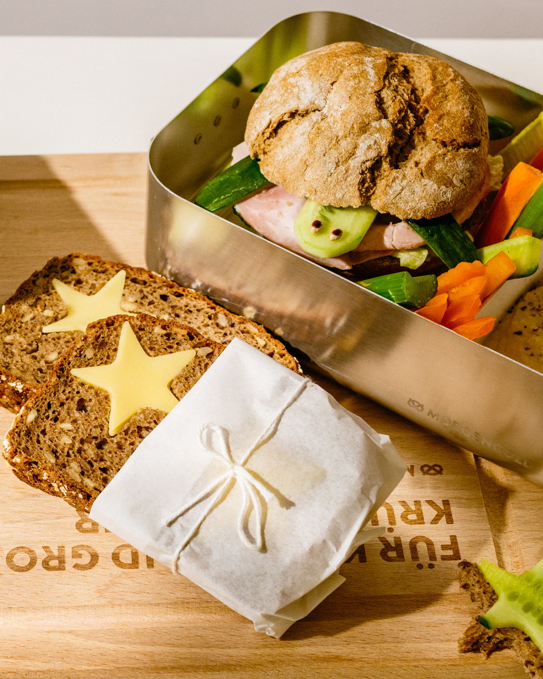 Vollkornbrot mit Käse-Sternen und verpacktem Pausenbrot neben einer Brotdose mit Brötchen, Rohkost und Dip.