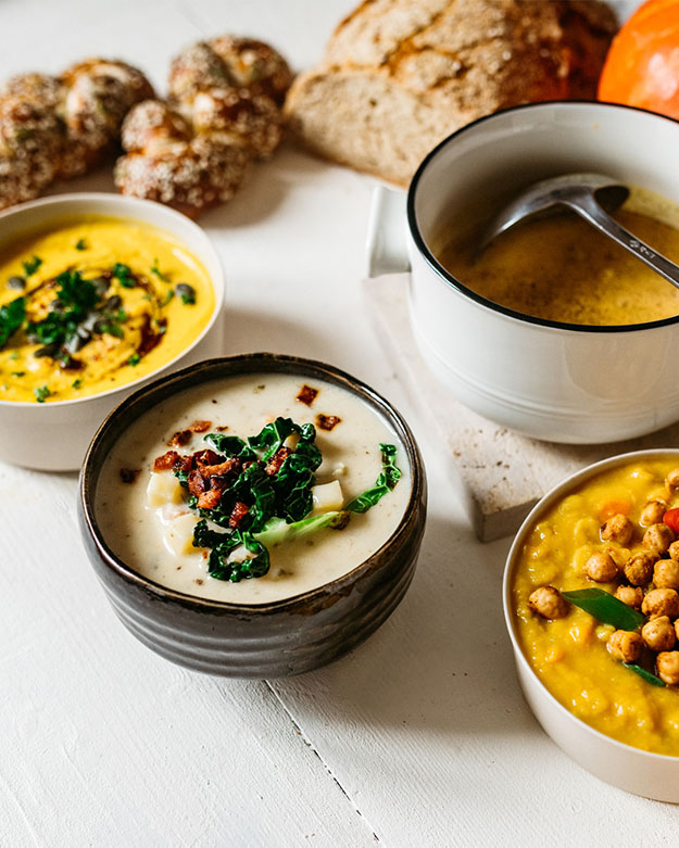 Vier herbstliche Suppenvariationen in Schalen angerichtet, darunter Kürbis-, Kartoffel-Grünkohl- und Linsensuppe mit Brot und Laugengebäck auf weißem Holztisch.