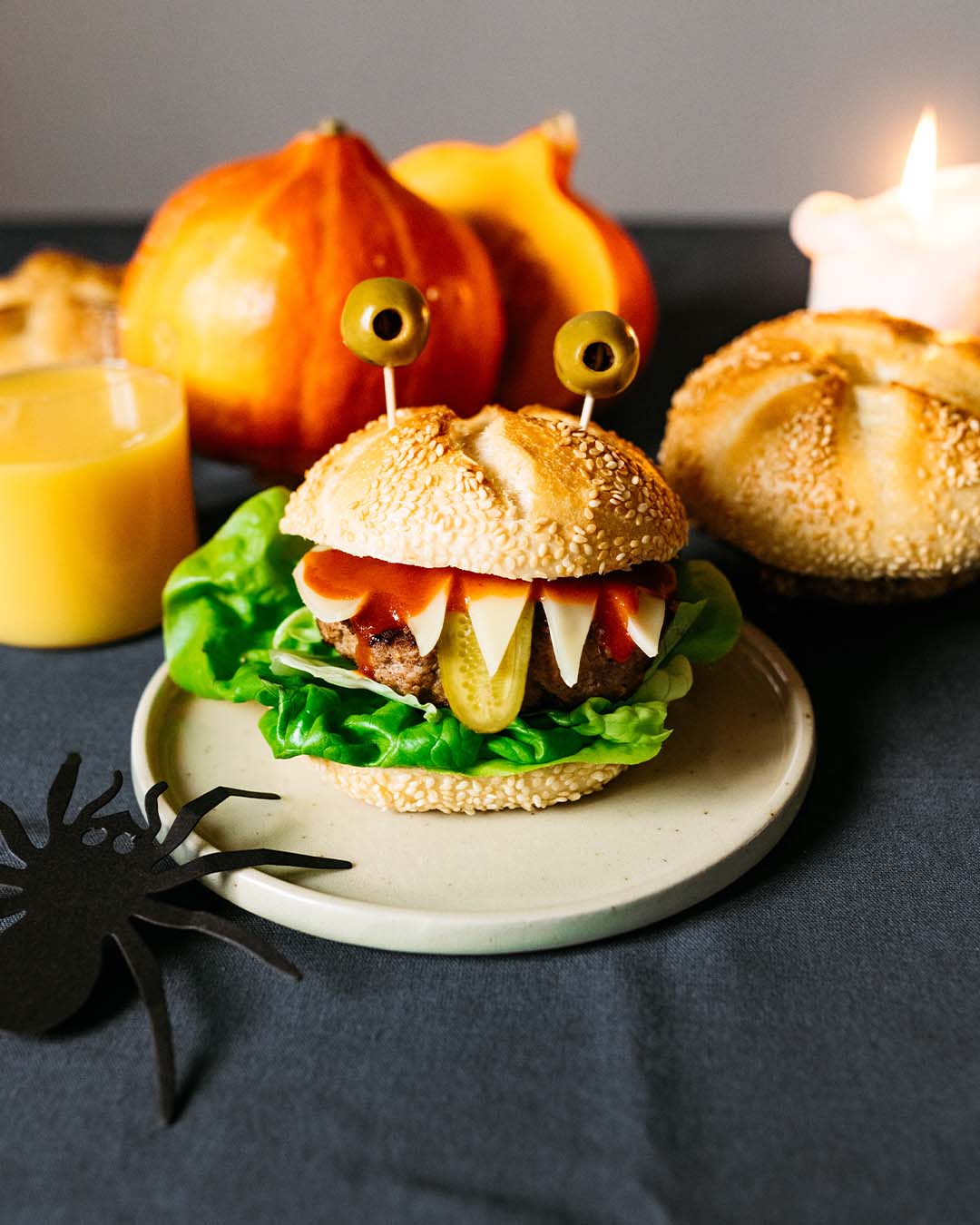 Halloween-Burger mit lustigem Monster-Gesicht aus Sesambrötchen, Käsezähnen, eingelegter Gurke, Olivenaugen und Salat, dekoriert mit Kürbissen, Saft, Kerze und einer schwarzen Papierspinne.