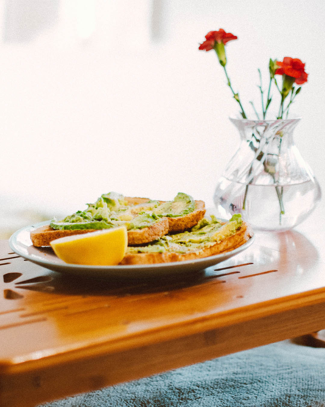 Frühstück im Bett mit Avocado-Toasts und Zitronenschnitz auf einem Holztischtablett, daneben eine Vase mit roten Nelken.