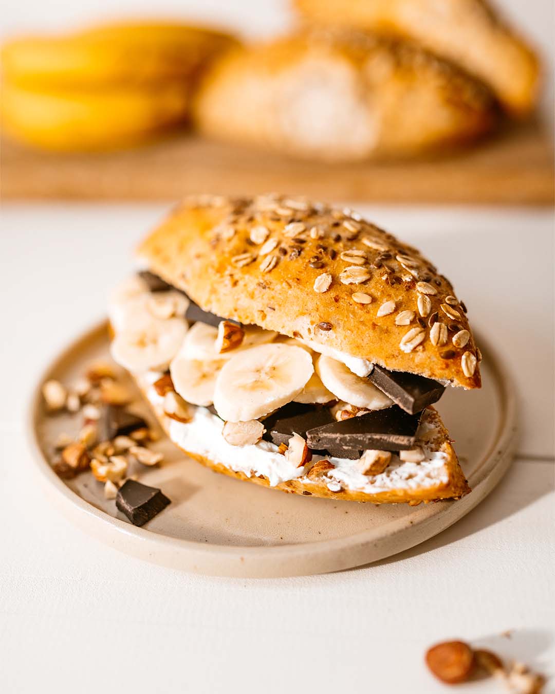 Körniges Brötchen mit Frischkäse, Bananenscheiben, dunkler Schokolade und gehackten Nüssen, angerichtet auf einem hellen Teller.