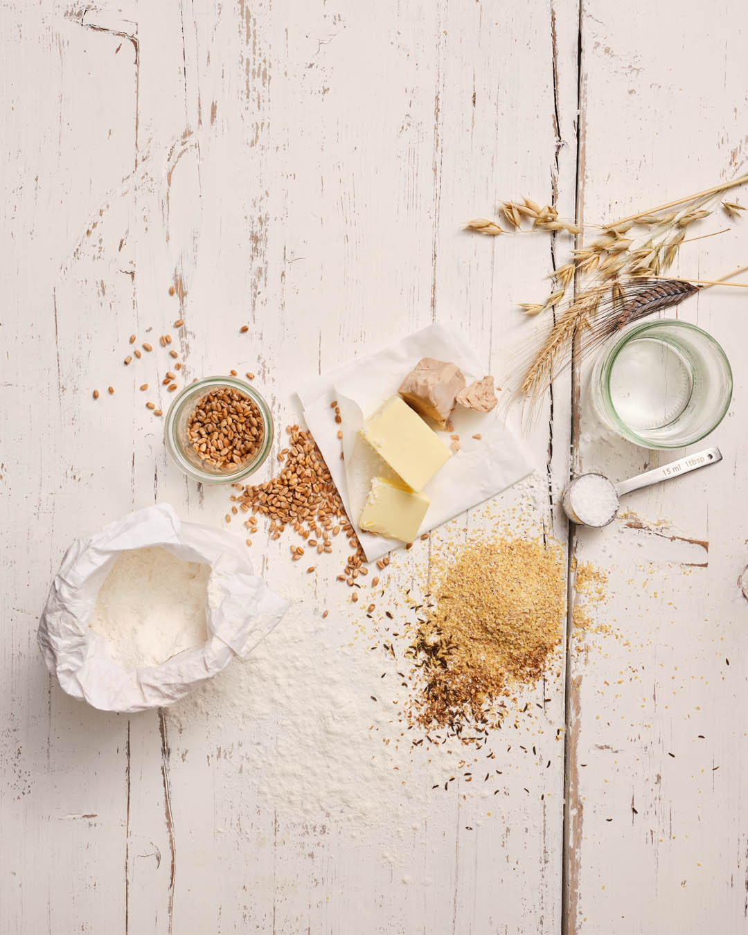 Verschiedene Brot-Zutaten auf einem weißen, rustikalen Holztisch – darunter Mehl, Butter, Hefe, Wasser, Körner, Getreideähren und ein Glas mit Weizenkörnern.