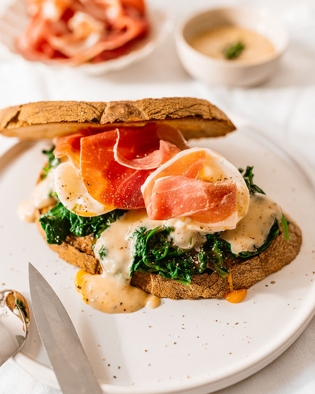 Rustikales Brot belegt mit gedünstetem Spinat, pochierten Eiern, Schinkenscheiben und cremiger Sauce, serviert auf einem weißen Teller.