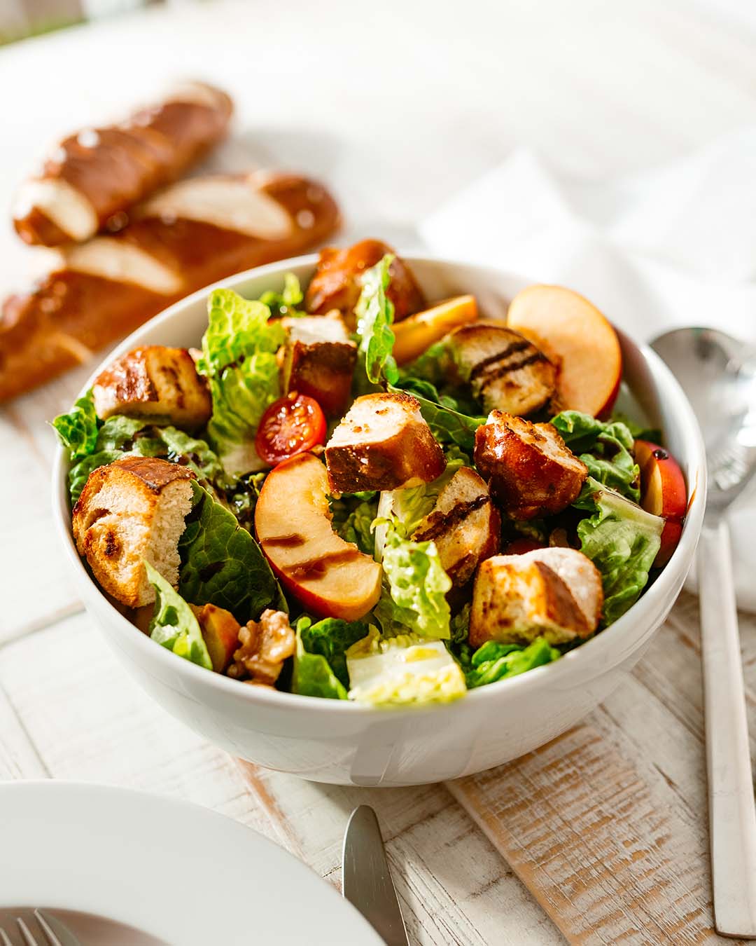 Romana-Salat mit Laugen-Croûtons, Pfirsichspalten und Kirschtomaten in einer weißen Schale, serviert mit Besteck auf Holztisch.
