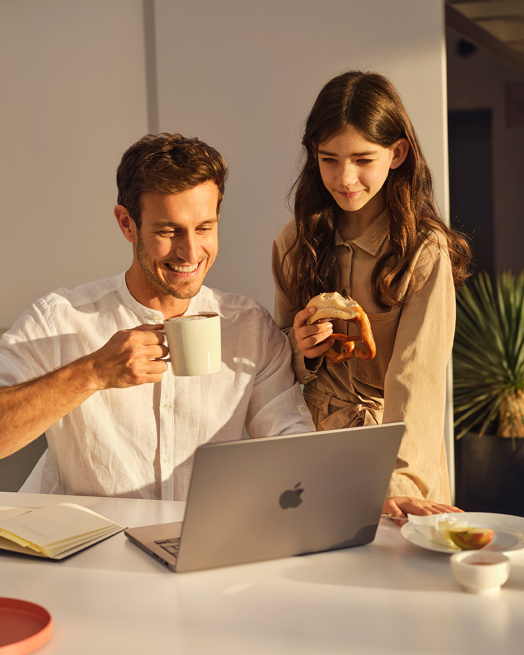 Vater trinkt Kaffee am Laptop, Tochter isst frische Brezel – gemeinsames Frühstück mit Morgengold Backwaren.