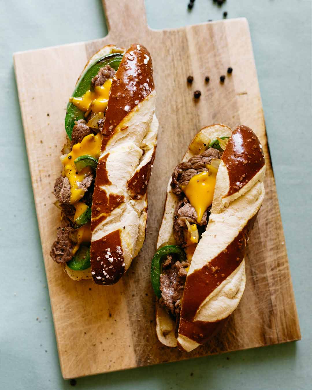 Zwei Philly Cheese Steak Sandwiches im Laugenbrötchen mit Rindfleisch, Cheddarkäse und grüner Paprika auf Holzbrett.