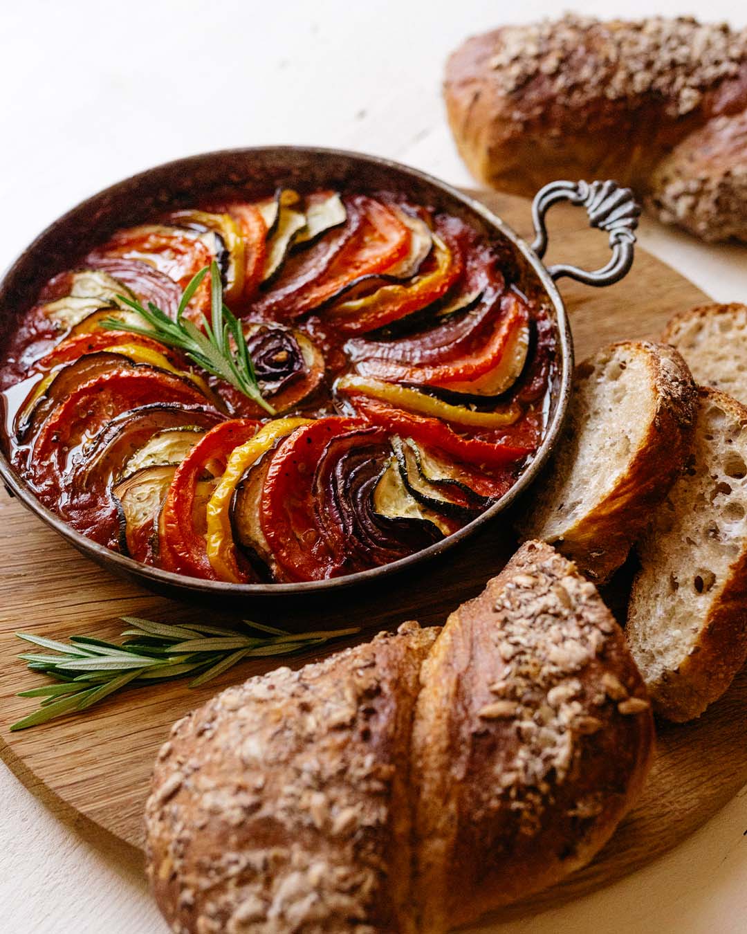 Ofenfrische Ratatouille mit buntem Gemüse in Scheiben, serviert mit knusprigem Körnerbrot auf Holzbrett.