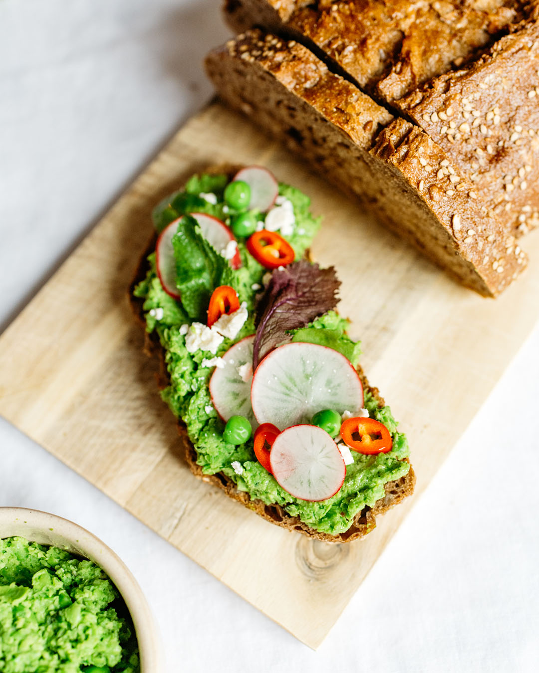 Frisches Vollkornbrot mit Erbsenpüree, Radieschen, Chili, Feta und Blattsalat auf Holzbrett serviert.