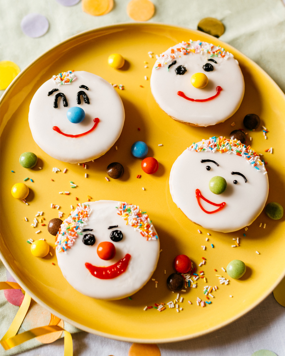Bunte Amerikaner mit Zuckerguss, Schokolinsen und Streuseln als lustige Gesichter auf gelbem Teller dekoriert.
