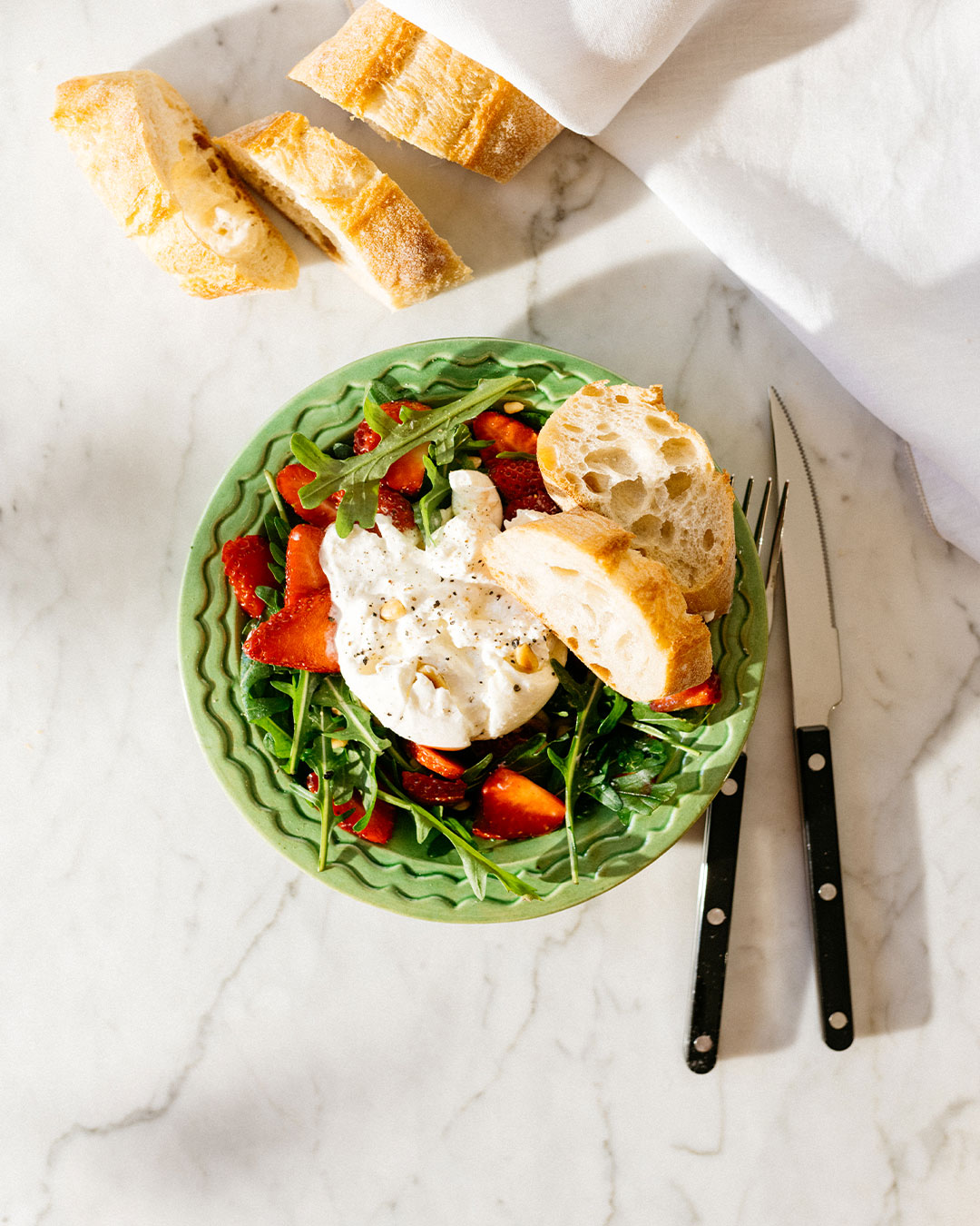 Frischer Erdbeersalat mit Burrata auf Rucola, serviert mit knusprigem Baguette auf grünem Teller.