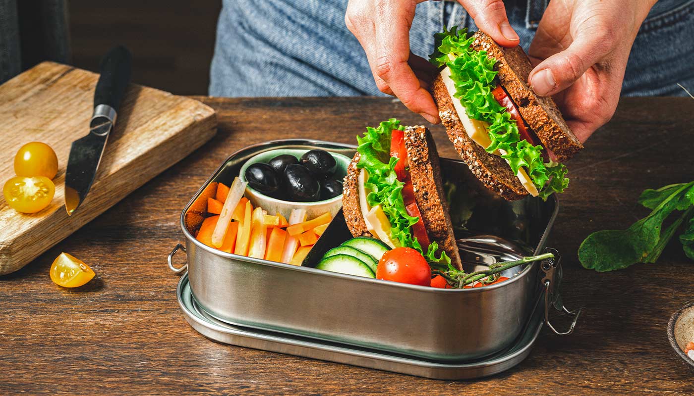 Vollkornsandwich mit Salat, Käse und Tomate in Lunchbox, dazu Gemüsesticks, Gurken und schwarze Oliven.