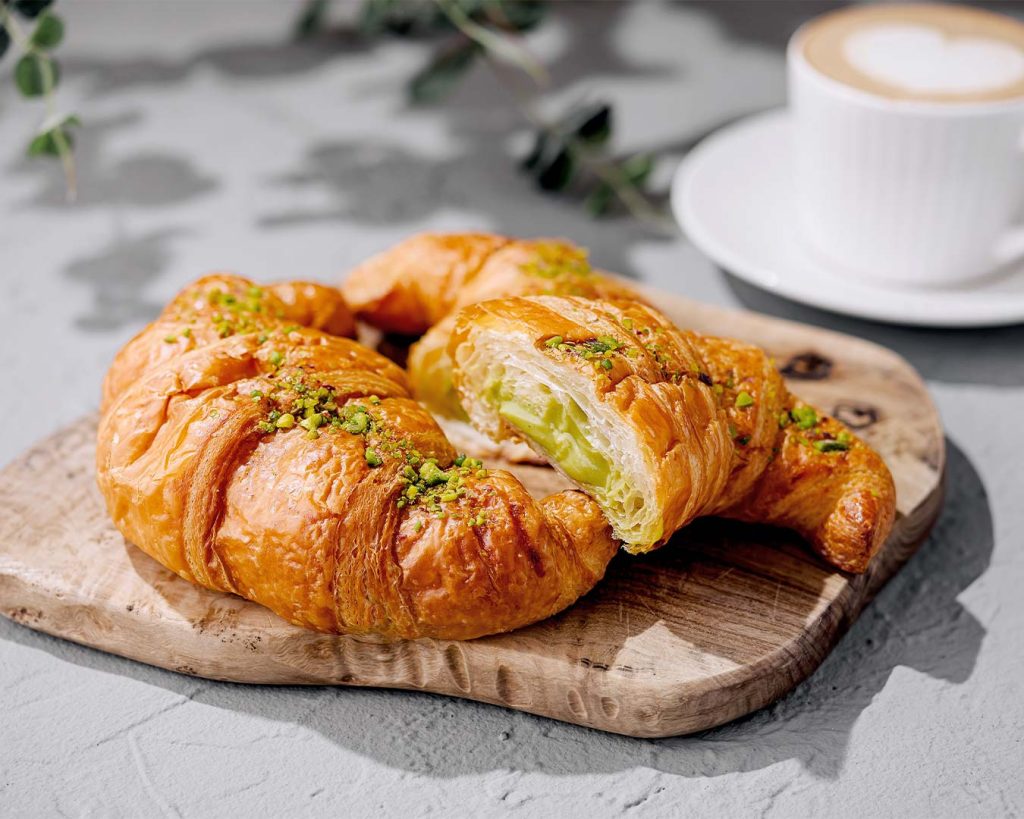 Aufgeschnittenes Croissant mit grüner Pistaziencreme-Füllung und gehackten Pistazien auf Holzbrett.